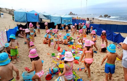Fotografia de cerca de quatro dezenas de crianças na praia. A areia está repleta de brinquedos. Ao fundo estão seis barracas de praia. Vê-se o mar e, mais longe ainda, a Capela do Senhor da Pedra (junto ao mar). Todos os rostos das crianças foram desfocados.