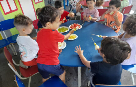 Oito crianças sentadas à volta de uma mesa azul redonda. Na mesa, dentro dos pratos, estão vários frutos com os quais as crianças estão a fazer espetadas de fruta. Todos os rostos das crianças foram desfocados.