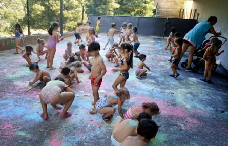 Perto de duas dezenas de crianças, a usar fato de banho, brincam em espaço exterior com pó de várias cores. Todos os rostos das crianças foram desfocados.