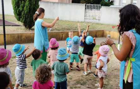 Em jardim exterior estão 13 crianças e duas pessoas adultas a fazerem bolas de sabão. Algumas estão a perseguir as bolas de sabão. Todos os rostos das crianças foram desfocados.