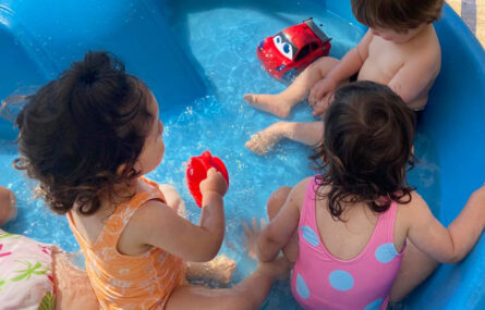 Três crianças, ainda muito jovens, sentadas dentro de uma pequena piscina insuflável cheia de água.