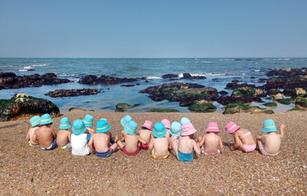 Fotografia à beira-mar. Em primeiro plano a areia da praia. Depois estão sentadas (de costas) 18 crianças, todas com chapéu de sol. O mar, estando em maré baixa, apresenta várias rochas repletas de algas.