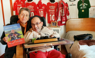 Fotografia de duas pessoas em espaço interior (quarto). Na imagem o atleta de Boccia da APPC, Armando Costa, acompanhado pela antiga atleta Rosa Mota. Rosa Mota segura na mão direita o livro “Armando Costa – fotobiografia de um vencedor”. Ao fundo, na parede, estão penduradas sete camisolas – sendo que cinco delas são do Sport Lisboa e Benfica. No canto inferior direito está sobreposto o logótipo do “European Para Championships 2023 – Rotterdam”.