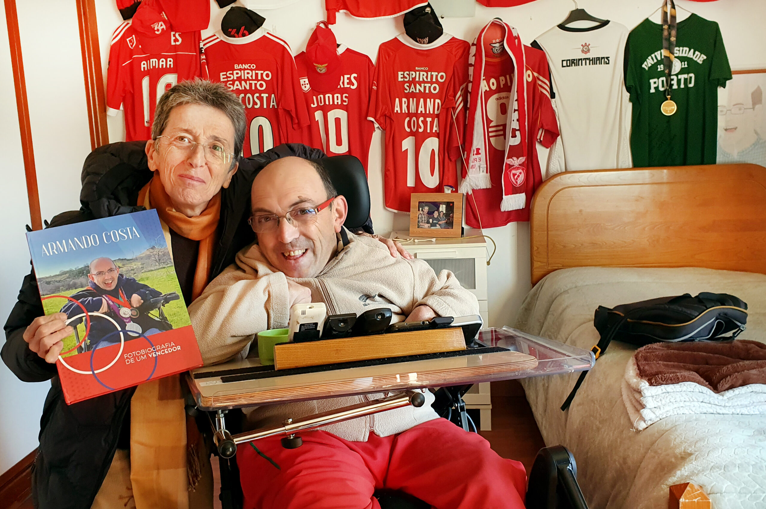 Fotografia de duas pessoas em espaço interior (quarto). Na imagem o atleta de Boccia da APPC, Armando Costa, acompanhado pela antiga atleta Rosa Mota. Rosa Mota segura na mão direita o livro “Armando Costa – fotobiografia de um vencedor”. Ao fundo, na parede, estão penduradas sete camisolas – sendo que cinco delas são do Sport Lisboa e Benfica. No canto inferior direito está sobreposto o logótipo do “European Para Championships 2023 – Rotterdam”.
