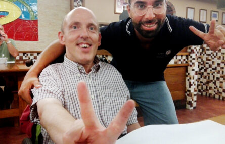 Junto de uma mesa duas pessoas sorridentes, com os dedos da mão, fazem (para a fotografia) um “sinal de vê” de Vitória.