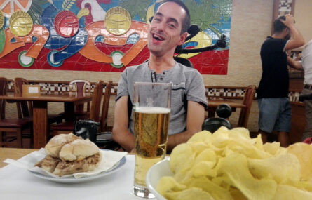 Uma pessoa sentada junto de uma mesa sorri para a fotografia. À sua frente, na mesa, o que aparenta ser uma sandes de leitão, um copo com cerveja e um prato cheio de batatas fritas. Ao fundo duas mesas vazias. E, na parede, um trabalho artístico em azulejo colorido.