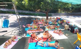 Grupo de cerca de duas dezenas de crianças em fato de banho deitadas em toalhas em jardim exterior. Estão à sombra de uma árvore. Do lado direito algumas mochilas e material diverso espalhado.