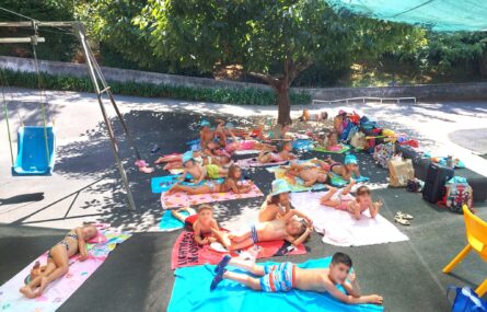 Grupo de cerca de duas dezenas de crianças em fato de banho deitadas em toalhas em jardim exterior. Estão à sombra de uma árvore. Do lado direito algumas mochilas e material diverso espalhado.