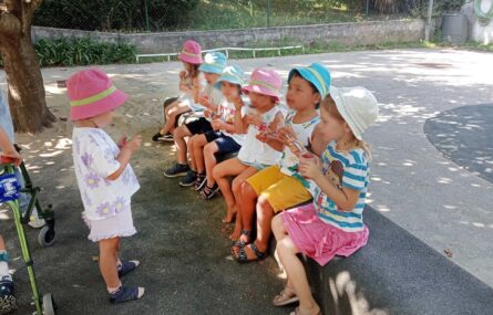 Seis crianças sentadas a comer algo (em espaço exterior, à sombra de uma árvore). De pé, à frente dessas seis crianças, está outra também a provar o que aparenta ser um gelado.