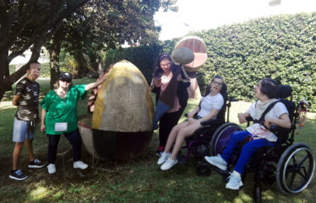 Grupo de cinco pessoas em jardim exterior – junto a algo que será uma escultura, em pedra, com o formato de um ovo. Ao fundo duas árvores e uma sebe.
