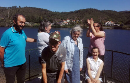 Grupo de seis pessoas, sorridentes, na margem de um rio. Do outro lado do rio algumas casas, montes e espaços verdes.