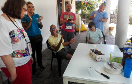 Seis pessoas junto de uma mesa na qual está pousado um bolo de aniversário com duas velas acesas. Em cima da mesa estão, também, alguns pratos, copos, guardanapos e uma garrafa de sumo.