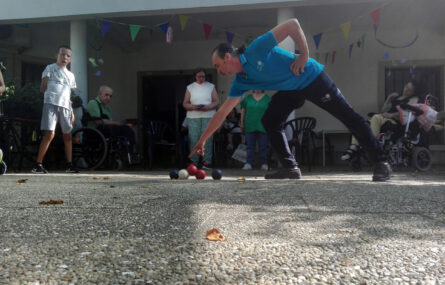 Várias pessoas assistem a um jogo de Boccia. Em primeiro plano está uma pessoa que aponta para uma das bolas de Boccia pousadas no chão.