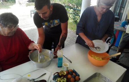 Três pessoas cozinham o que aparenta vir a ser um bolo. Em cima da mesa estão ingredientes e utensílios de preparação. Ao fundo vê-se parte de um jardim.