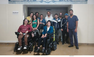 Grupo de pessoas junto de um acesso a espaço interior. Duas das pessoas usam cadeira de rodas.