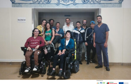 Grupo de pessoas junto de um acesso a espaço interior. Duas das pessoas usam cadeira de rodas.