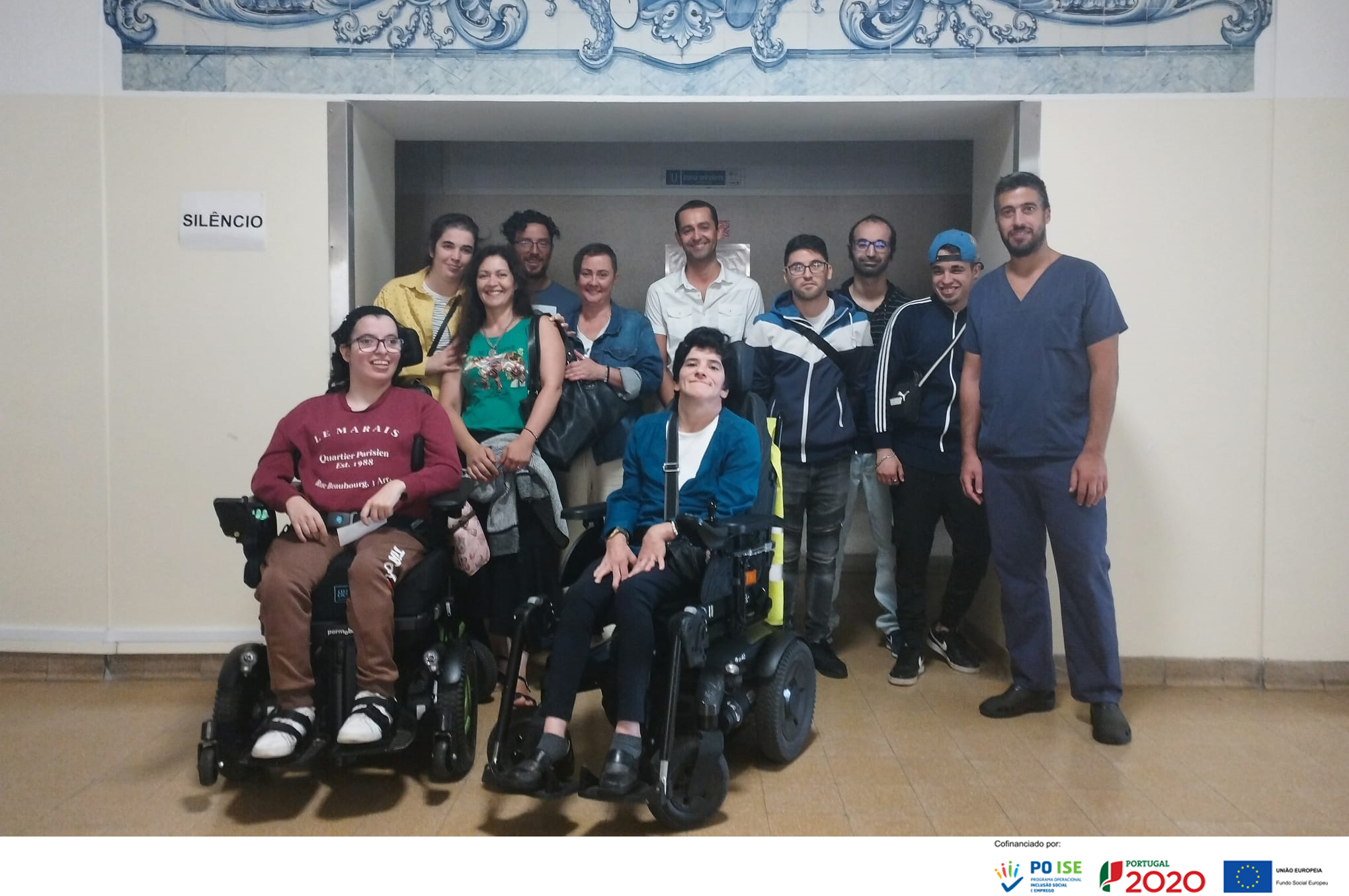 Grupo de pessoas junto de um acesso a espaço interior. Duas das pessoas usam cadeira de rodas.