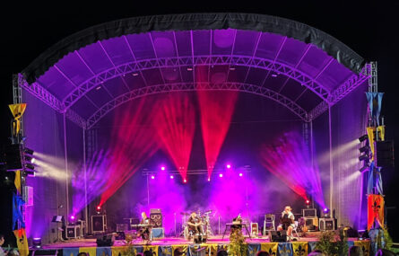 Os quatro elementos da banda appSound em palco, durante a atuação. Palco com iluminação essencialmente em tons de roxo e vermelho. Algumas bandeiras (ou estandartes) estão pendurados nas zonas laterais do palco.