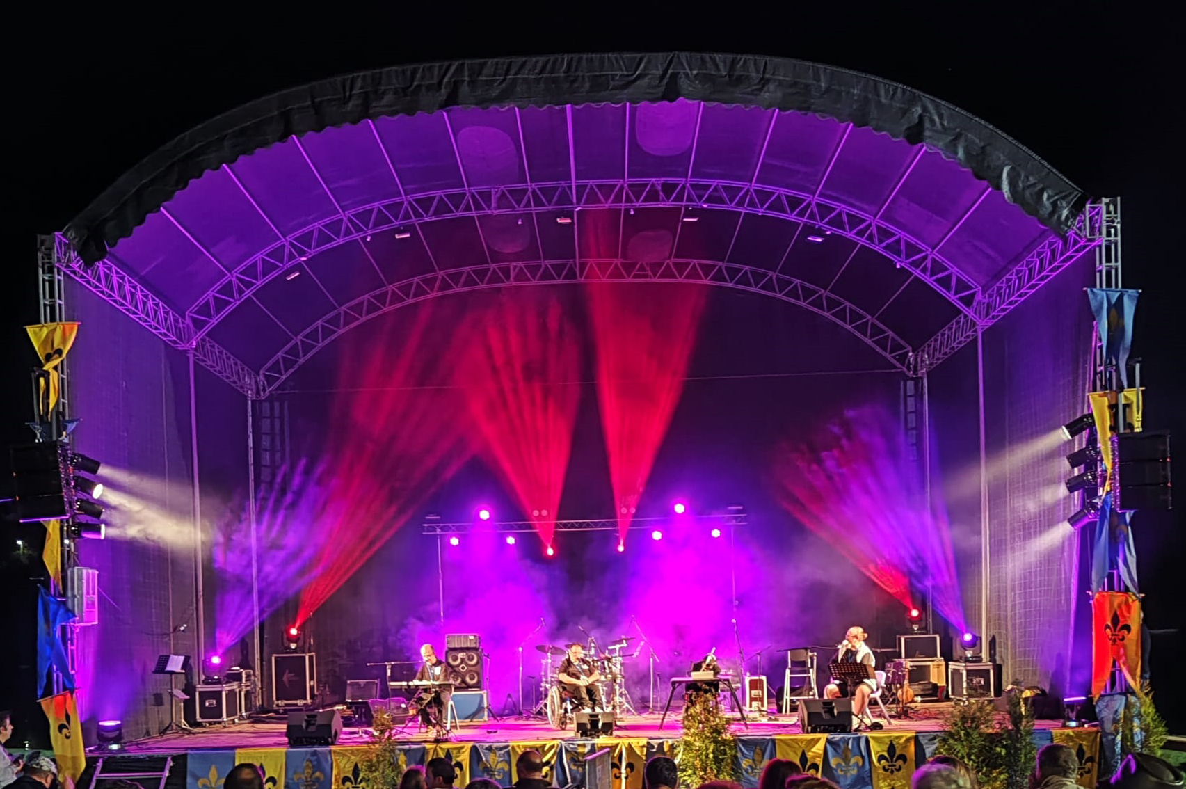 Os quatro elementos da banda appSound em palco, durante a atuação. Palco com iluminação essencialmente em tons de roxo e vermelho. Algumas bandeiras (ou estandartes) estão pendurados nas zonas laterais do palco.