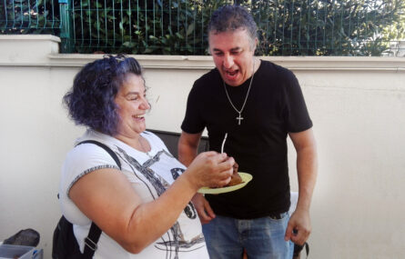 Fotografia de duas pessoas em espaço exterior (junto de um muro). Uma das pessoas, sorridente, segura o que aparenta ser uma vela de aniversário e um bolo. A outra pessoa estará prestes a apagar a vela.