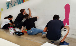 Fotografia de quatro jovens (sentados ou ajoelhados) durante o início da pintura de um mural. A parede branca começa já a ter algumas zonas com manchas rosa e azul.