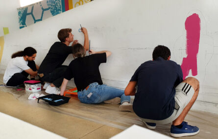 Fotografia de quatro jovens (sentados ou ajoelhados) durante o início da pintura de um mural. A parede branca começa já a ter algumas zonas com manchas rosa e azul.