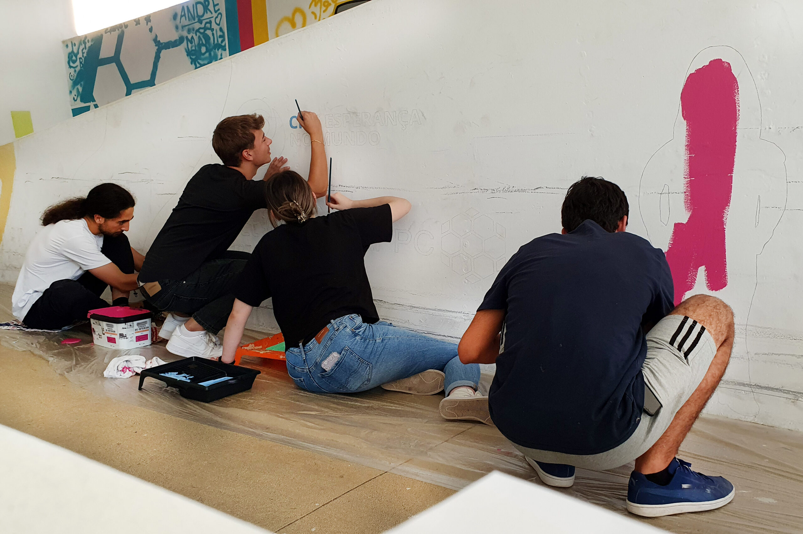 Fotografia de quatro jovens (sentados ou ajoelhados) durante o início da pintura de um mural. A parede branca começa já a ter algumas zonas com manchas rosa e azul.