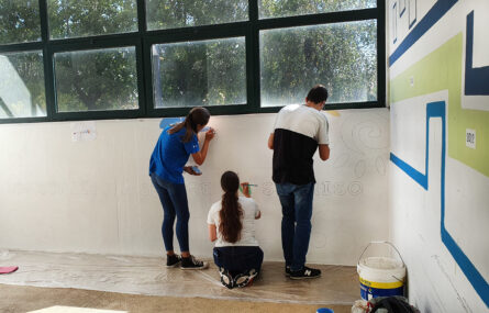 Fotografia de três jovens de costas na fase inicial de pintura de um mural. No chão estão pousados alguns materiais de pintura.