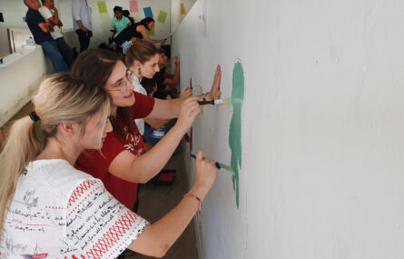 Perspectiva lateral de várias pessoas a pintar o mural. Em primeiro plano duas jovens pintam uma área de verde.