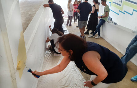 Imagem de várias pessoas a pintar um mural. Em primeiro plano uma senhora usa um rolo para pintar uma área extensa com tonalidade amarela. Mais ao fundo estão vários jovens a conversar e assistir.