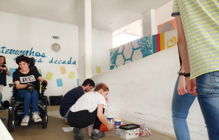 Dois jovens, ajoelhados, preparam materiais para a pintura do moral. São ainda parcialmente visíveis pessoas em ambos os lados da imagem. Ao centro uma pessoa em cadeira de rodas sorri enquanto aparenta avaliar a evolução das pinturas.