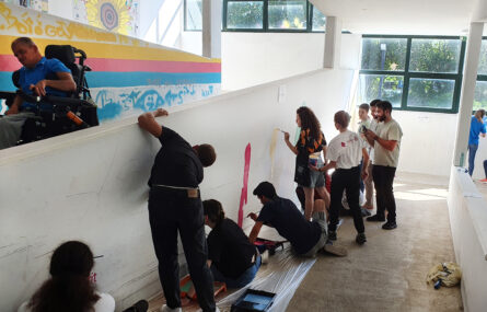 Perspetiva diagonal de uma dezena de jovens durante a pintura de um mural. A parede (branca) do mural apresenta já várias zonas coloridas. Vê-se ainda, em contraluz, uma pessoa a passar em cadeira de rodas.