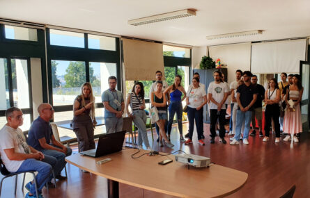 Imagem de sala ampla com perto de duas dezenas de jovens que assistem a uma apresentação. À esquerda estão dois representantes da APPC (os presidentes da Direção e do Conselho Fiscal). Em primeiro plano uma mesa com um computador e um projetor.