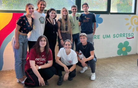 Fotografia de nove jovens junto de um dos murais pintados. Na parede (agora já colorida) consegue-se ler parcialmente uma mensagem escrita – que termina com a palavra “Sorriso”.
