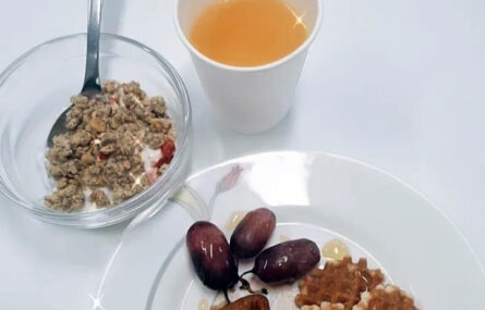Um copo com o que aparenta ser sumo de laranja. Ao lado está uma taça com cereais e iogurte e, mais abaixo, um prato com uvas, bolachas e uma fatia de bolo.