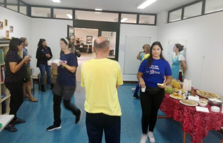 Fotografia com nove pessoas em sala interior. Aparentam estar a conversar e algumas seguram copos e algo para comer nas mãos. À direita vê-se uma mesa recheada de comidas e bebidas.