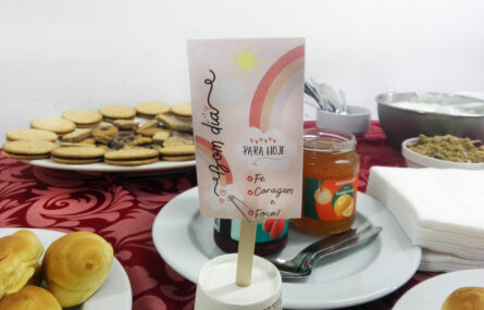 Fotografia de uma mesa com alguns produtos comestíveis (bolachas, biscoitos, compotas e croissants) bem como com alguns utensílios (pratos, talheres e guardanapos). Ao centro, em destaque, pequeno postal com a mensagem “Bom dia” e o menu do dia – composto por fé, coragem e foco!