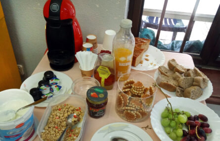 Imagem de uma mesa com inúmeros produtos comestíveis, sumos e máquina e pastilhas de café. Surgem ainda vários pratos, talheres e taças.