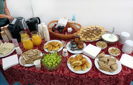 Fotografia de uma mesa com alguns produtos comestíveis (bolachas, biscoitos, queijo, cereais, compotas, uvas e croissants) bem como com alguns utensílios (pratos, talheres e guardanapos). À esquerda vê-se parcialmente alguém a usar uma máquina de café.