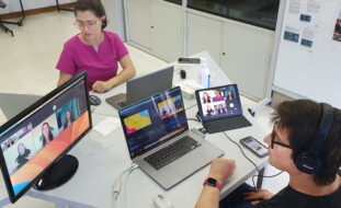 Imagem em plano superior de duas pessoas sentadas junto de mesa octogonal. Na referida mesa estão dois computadores portáteis em utilização, um outro ecrã de computador e ainda um tablet e telemóvel. Nos ecrãs visíveis pode-se acompanhar a transmissão de uma das emissões do webinar (vendo-se ainda as pessoas que participaram no painel de debate de 28 de setembro).