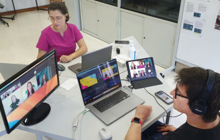 Imagem em plano superior de duas pessoas sentadas junto de mesa octogonal. Na referida mesa estão dois computadores portáteis em utilização, um outro ecrã de computador e ainda um tablet e telemóvel. Nos ecrãs visíveis pode-se acompanhar a transmissão de uma das emissões do webinar (vendo-se ainda as pessoas que participaram no painel de debate de 28 de setembro).