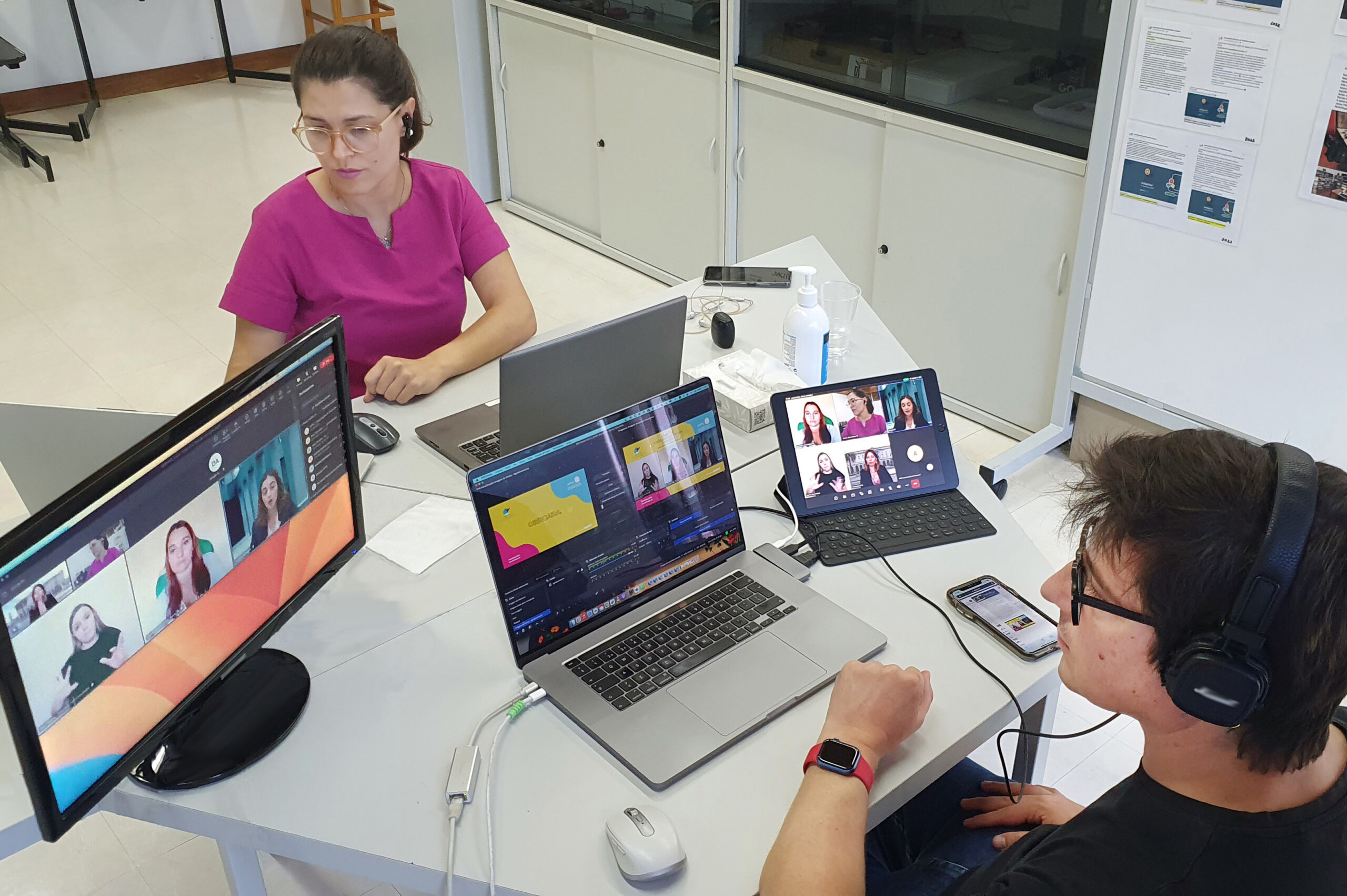 Imagem em plano superior de duas pessoas sentadas junto de mesa octogonal. Na referida mesa estão dois computadores portáteis em utilização, um outro ecrã de computador e ainda um tablet e telemóvel. Nos ecrãs visíveis pode-se acompanhar a transmissão de uma das emissões do webinar (vendo-se ainda as pessoas que participaram no painel de debate de 28 de setembro).