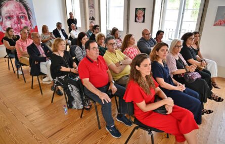 Fotografia de espaço interior com mais de duas dezenas de pessoas sentadas em várias filas de cadeiras. Ao fundo, na parede, uma pintura com um rosto. Na parede lateral mais algumas pinturas em quadros. Uma pessoa assiste de pé ao fundo da sala.