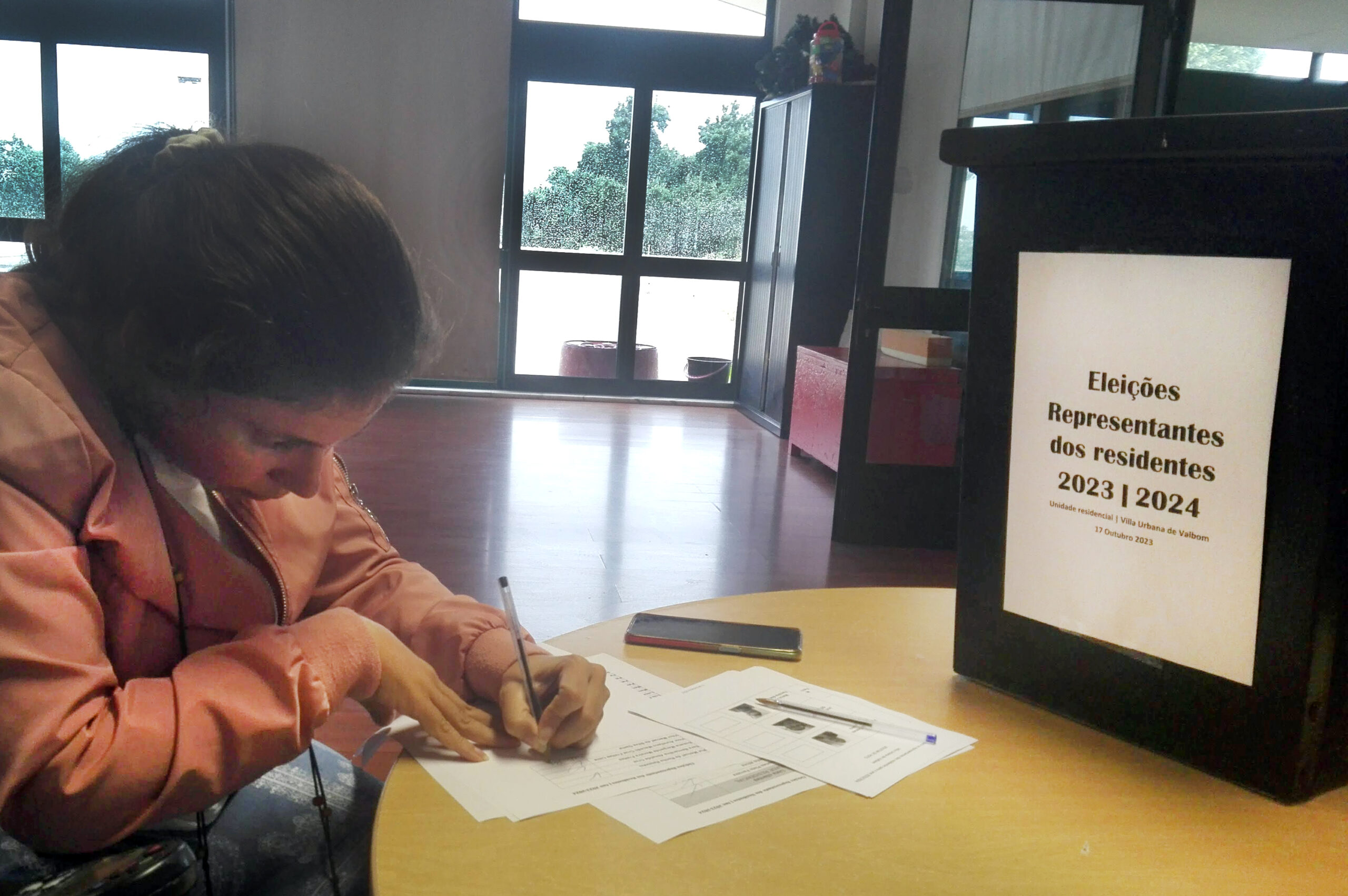 Pessoa a preencher boletim de voto pousado em cima de uma mesa. Também na mesa, mais à direita, está uma urna de votos com uma folha impressa onde se lê “Eleições Representantes dos Residentes 2023-2024”.