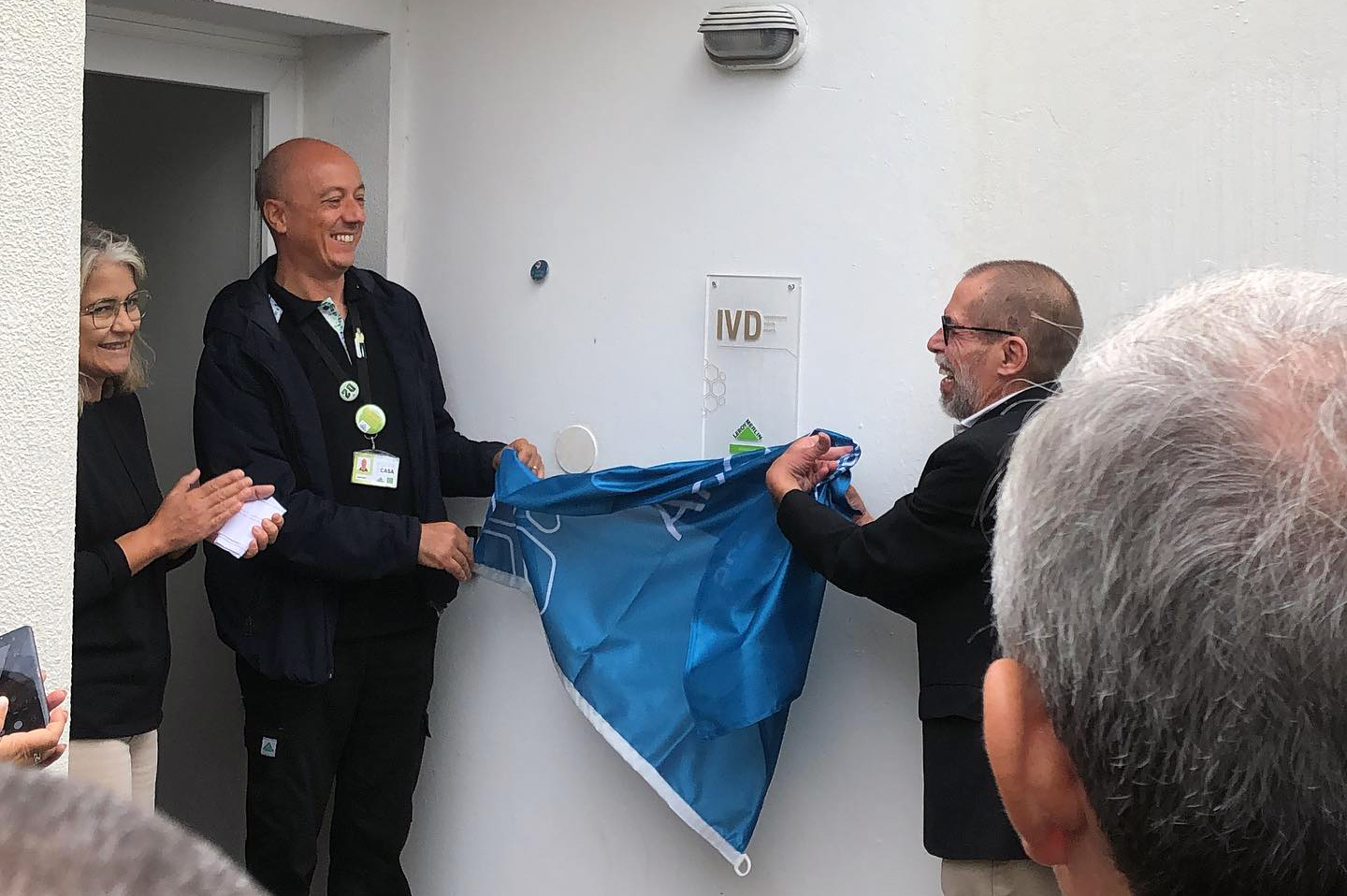 Duas pessoas retiram uma bandeira, da APPC, que cobria uma placa (com a indicação IVD). Mais à esquerda vê-se, parcialmente, uma terceira pessoa – igualmente sorridente.