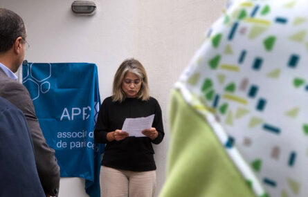 Uma pessoa lê o conteúdo de algo escrito numa folha de papel. Ao seu lado, nas costas, está uma bandeira azul da APPC que estará a tapar uma placa (ainda por descobrir).