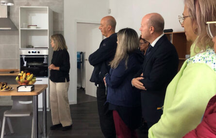 Seis pessoas dentro de espaço interior – em tudo semelhante a um pequeno apartamento. Ao fundo vê-se uma bancada e alguns eletrodomésticos de cozinha