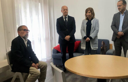 Três pessoas (à direita, em pé) aparentam ouvir o que uma quarta pessoa, sentada, à esquerda, estará a dizer. Junto à parede vê-se um sofá azul e, mais acima, um aparelho de ar condicionado. Em primeiro plano está uma mesa redonda.