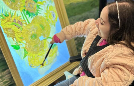 Uma pessoa utiliza um pincel num quadro interativo que simula uma pintura com várias flores de tom amarelo.