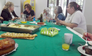 Cinco pessoas sentadas à mesa e a conversar. Em cima da mesa estão vários bolos, frutas laminadas, sumos, pratos e copos.